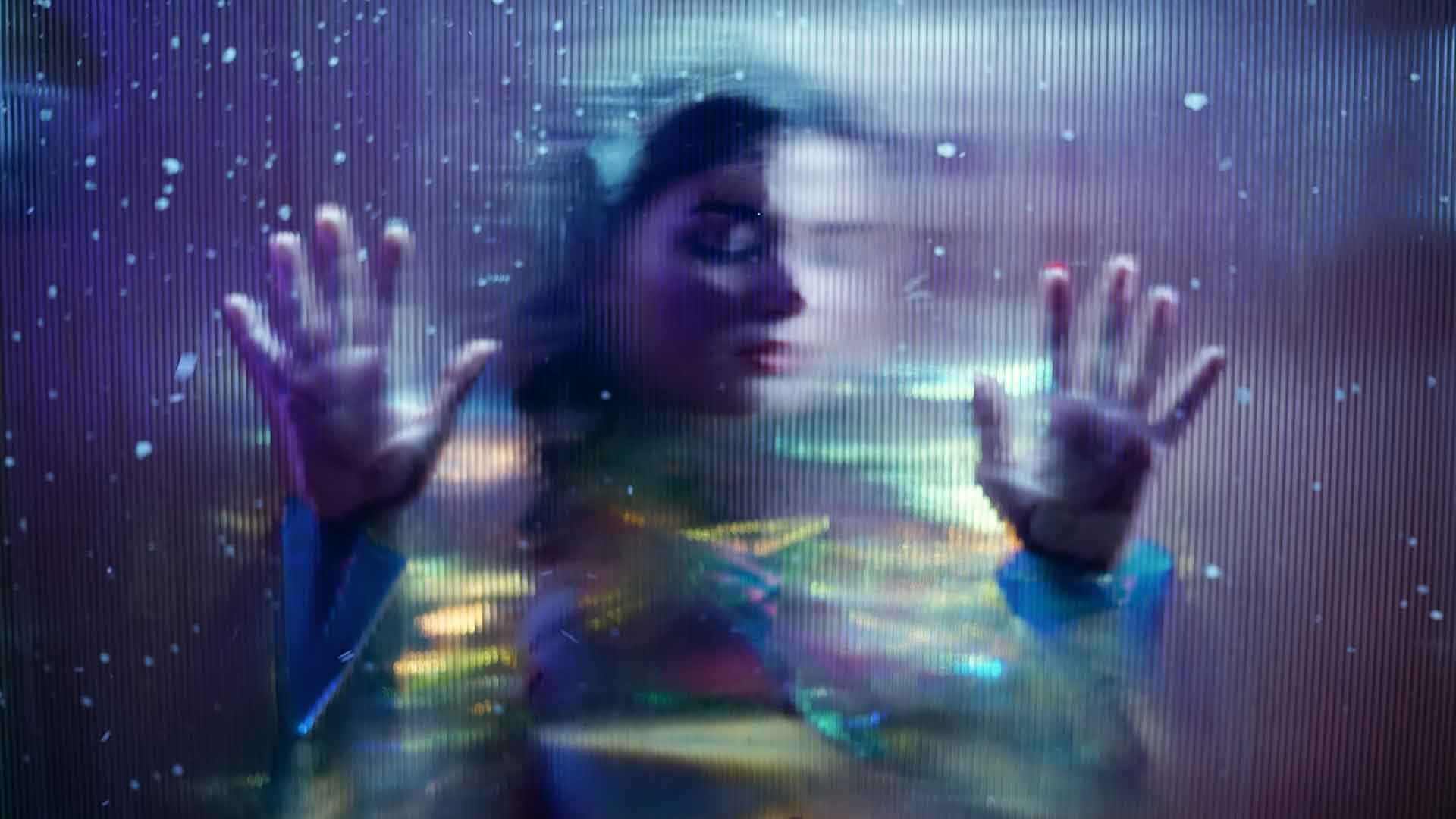Une femme est contre une vitre. Elle est entourée de couleurs froides