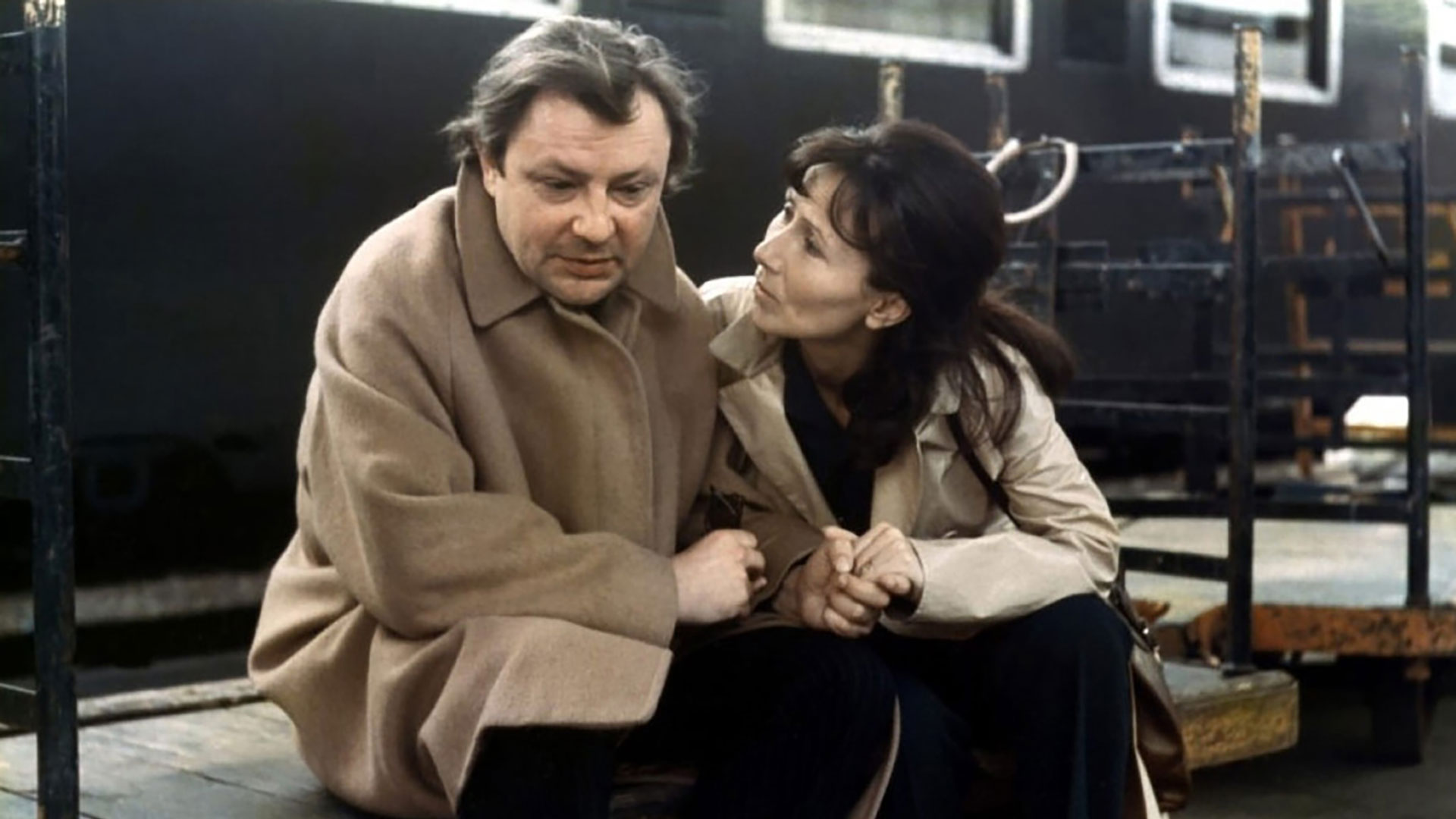 Un homme et une femme dans une gare sont assis sur un banc. L'homme est soucieux et la femme lui tient la main