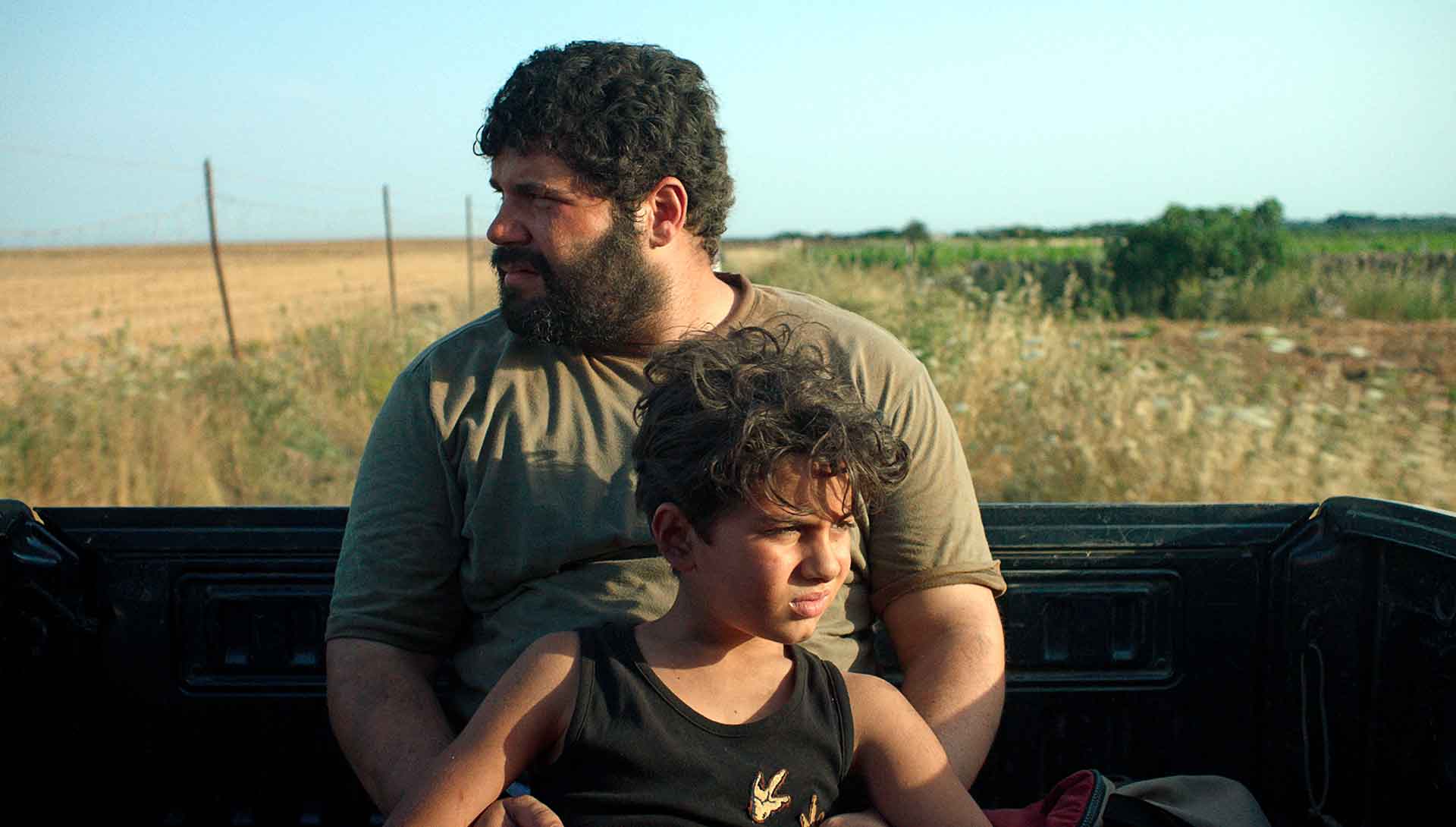 Un homme et un enfant sont assis à l'arrière d'un pick-up en pleine campagne. Extrait du film Una Promessa de Gianluca et Massimiliano De Serio.